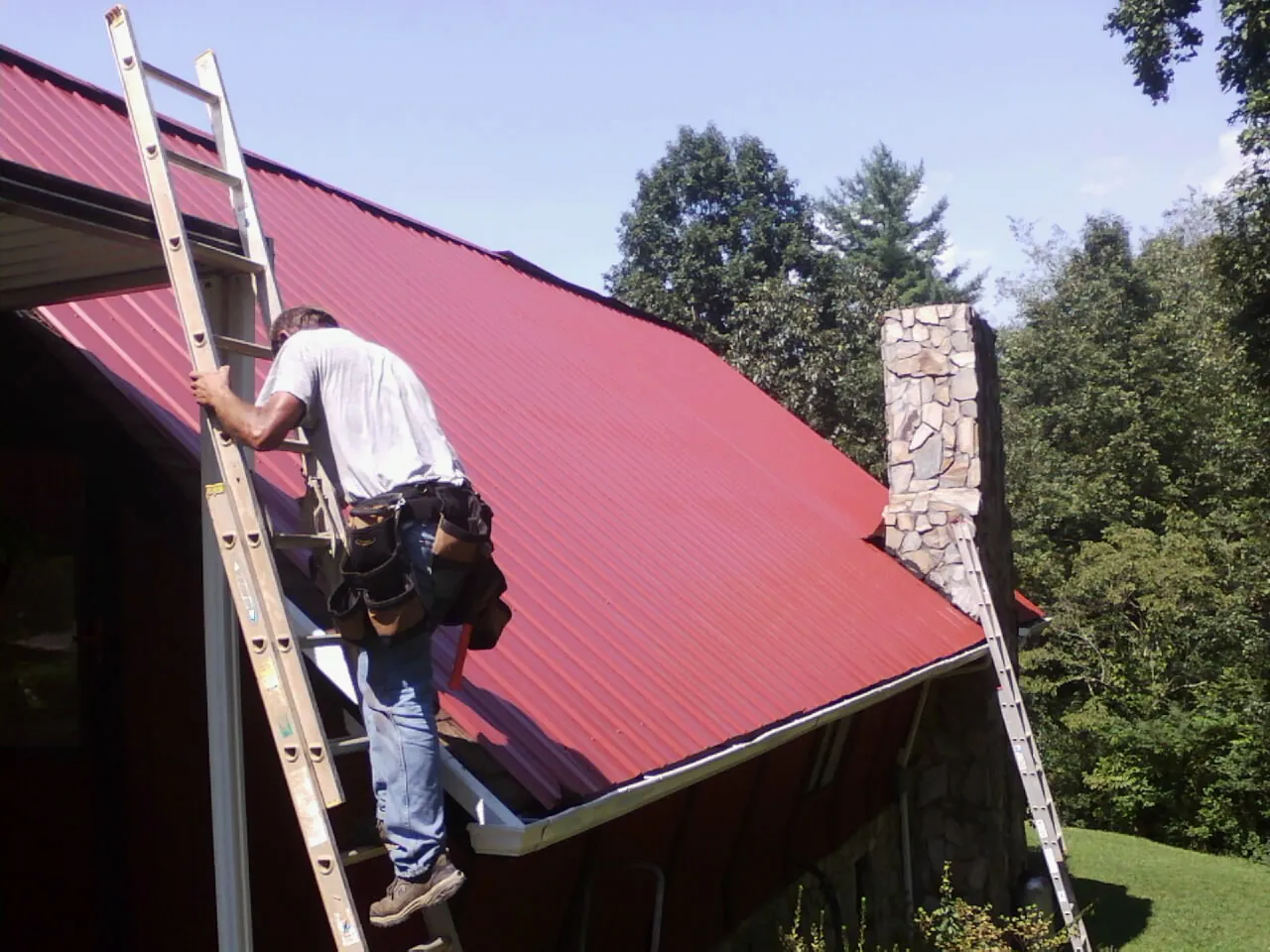 Roofer on a ladder beside a red metal roof and stone chimney — Soffit & Fascia Repair by Roofing Brothers in Mattaponi